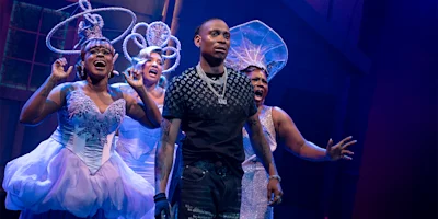 A man in casual clothes stands center stage as three women in elaborate white costumes and headpieces sing behind him under stage lighting.