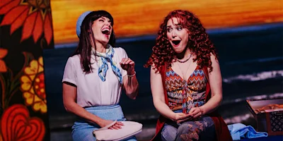 Two women sit next to each other on stage, both with wide smiles and surprised expressions, in colorful costumes with a vibrant background.