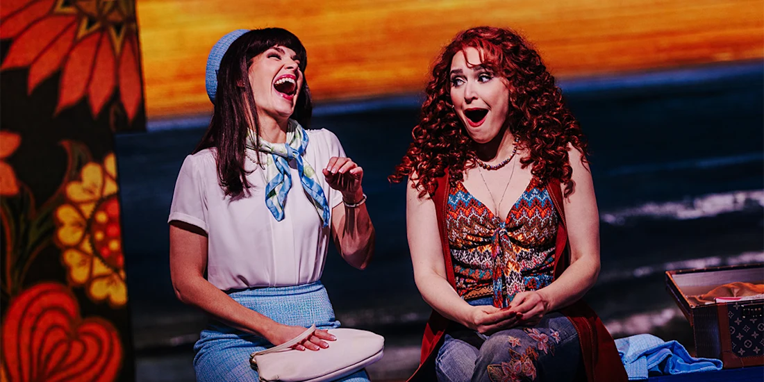 Two women sit next to each other on stage, both with wide smiles and surprised expressions, in colorful costumes with a vibrant background.