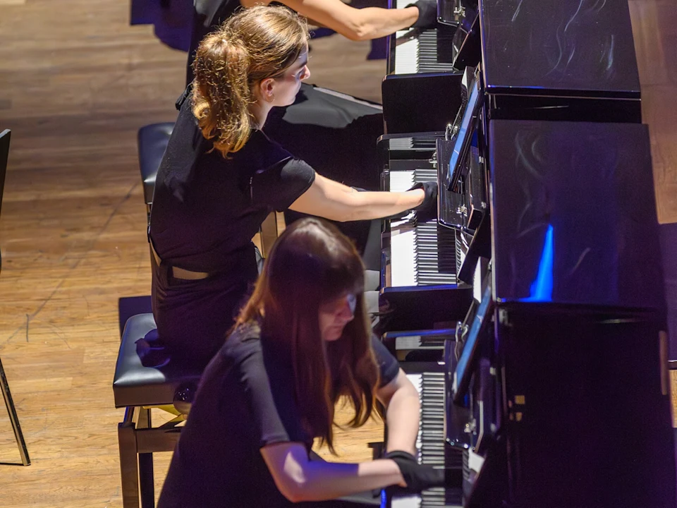 Two people in black clothing play upright pianos on a wooden stage, viewed from above.
