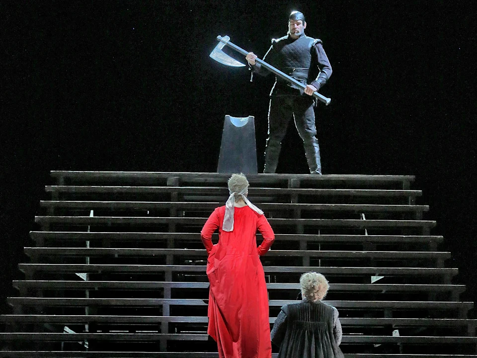 A person in black holds a large axe atop a staircase, facing two people below, one in red robes and the other in darker clothing, against a dark background.