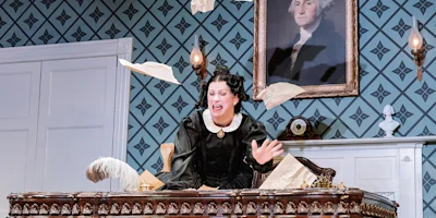 A woman in period costume sits at a desk with papers flying in the air, in a room with patterned wallpaper and a portrait of George Washington on the wall.