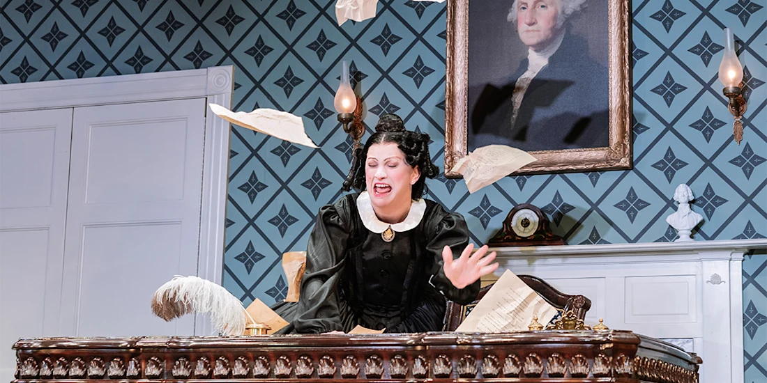 A woman in period costume sits at a desk with papers flying in the air, in a room with patterned wallpaper and a portrait of George Washington on the wall.