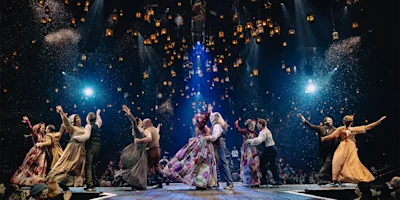 Actors in period costumes dance in pairs on a stage, surrounded by floating lanterns and illuminated by spotlights, with an audience seated around them.
