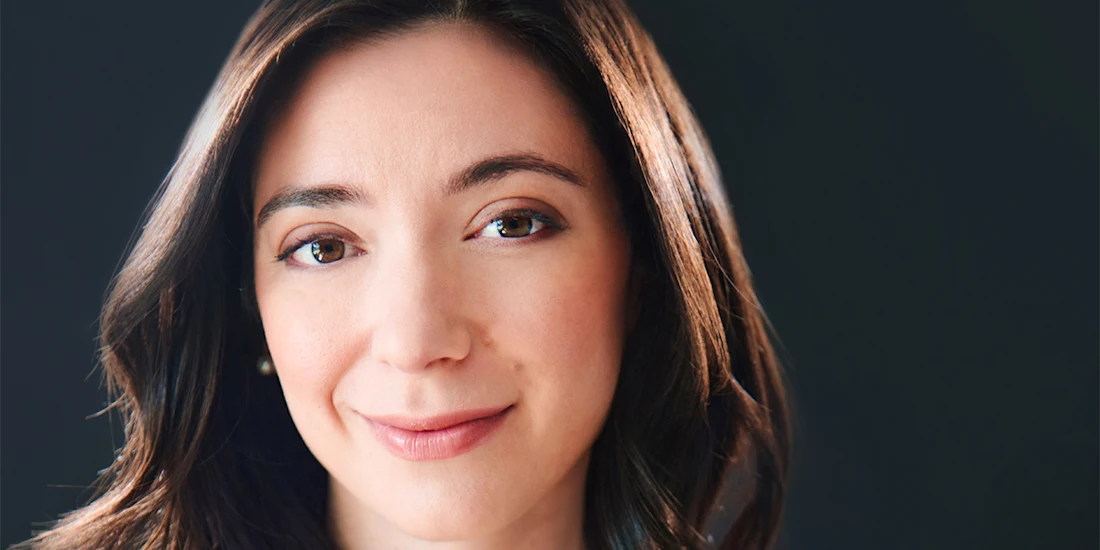 A woman with medium-length dark brown hair and light skin looks at the camera with a neutral expression against a dark background.