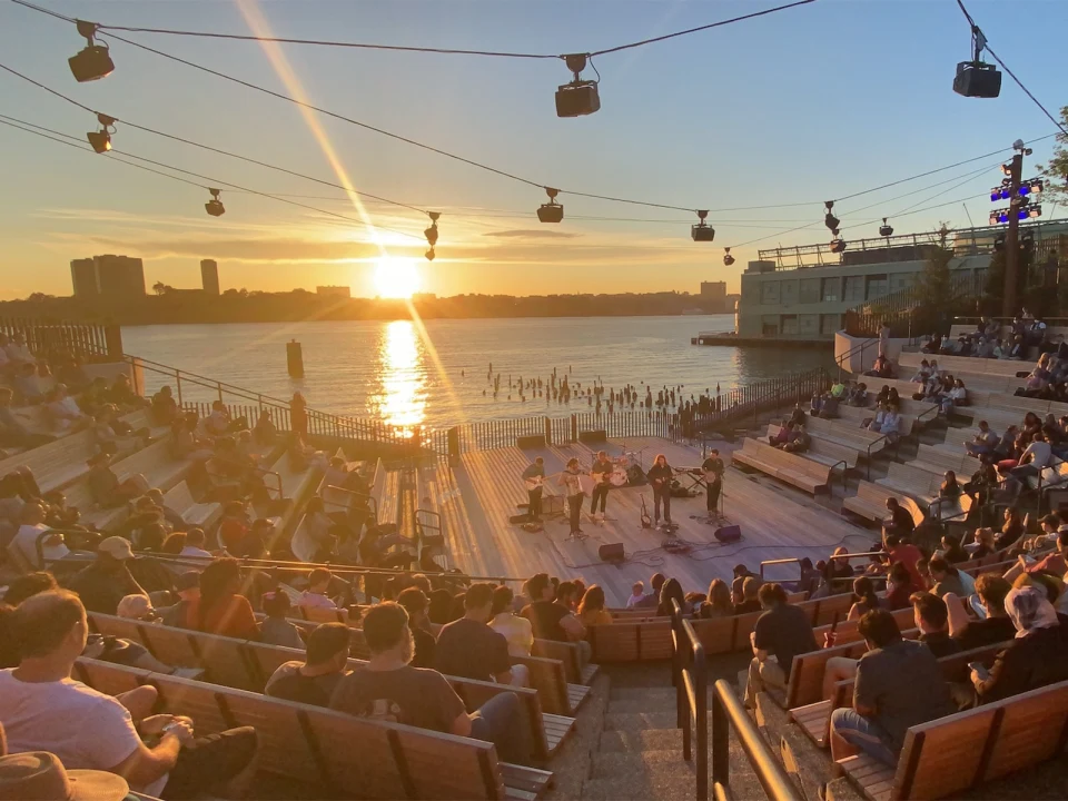 Sunset view over a river with people watching a live performance in an outdoor amphitheater, cable cars visible overhead.