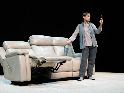 A person stands beside a light-colored reclining sofa, holding a phone and looking at the screen with a neutral expression.