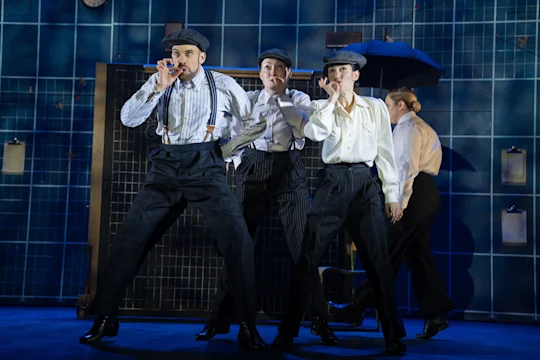 Three people in vintage-style outfits and flat caps dance and mime smoking cigars on a stage with a blue grid background and props. Another person stands in the background.