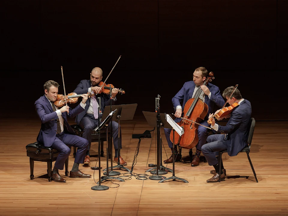 Four musicians in suits perform a string quartet on stage, each playing a different string instrument with sheet music stands and microphones in front of them.