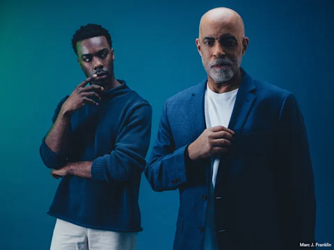 Two men stand against a blue background; one is older with a beard in a suit, the other is younger, wearing a sweater and appears to be thinking.