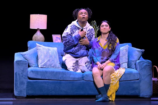 Two people are sitting on a blue couch on stage. One looks up with a surprised expression, while the other looks ahead. A lamp and framed photo are on a side table.