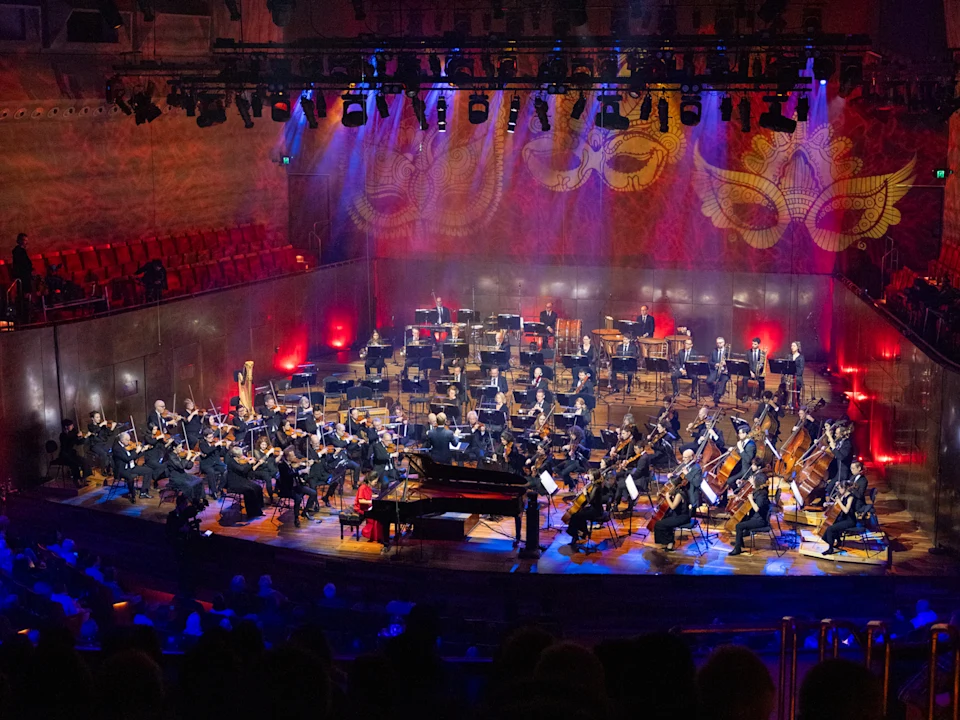 A full orchestra performs onstage in a large, well-lit concert hall with red and purple lighting and decorative patterns projected on the back wall.