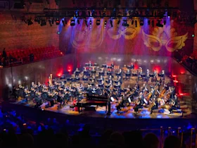 A full orchestra performs onstage in a large, well-lit concert hall with red and purple lighting and decorative patterns projected on the back wall.