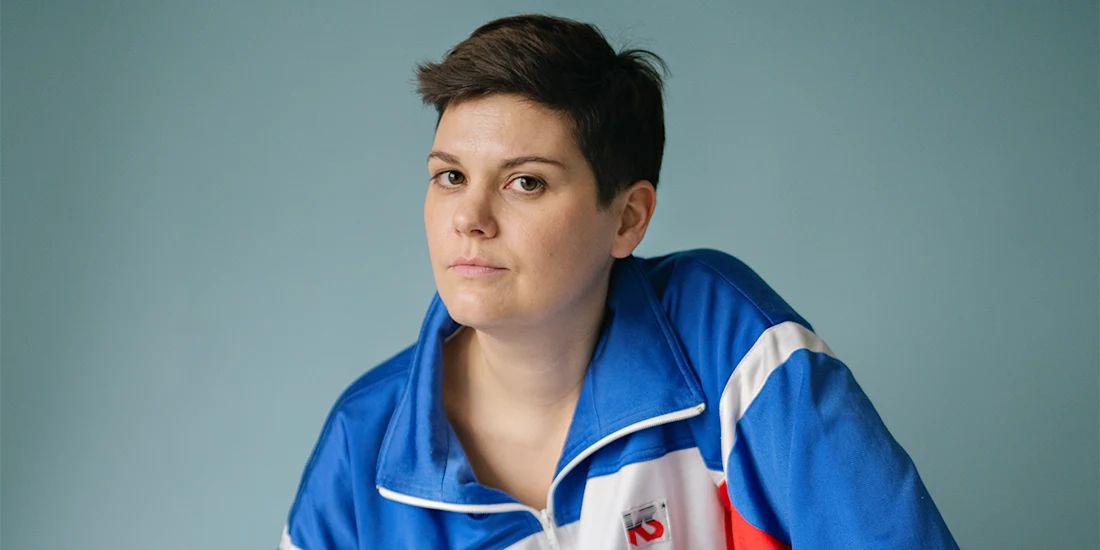 A person with short dark hair wearing a blue, white, and red jacket looks at the camera against a plain light blue background.