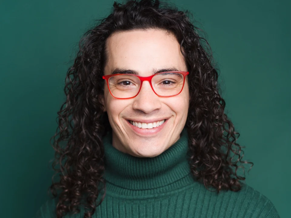 Person with long curly hair and red glasses smiling, wearing a green turtleneck.