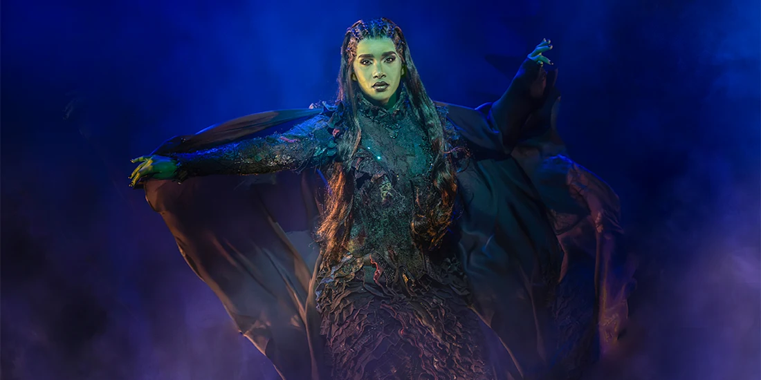 Performer in a dramatic, dark costume with outstretched arms stands on a smoky, blue-lit stage.