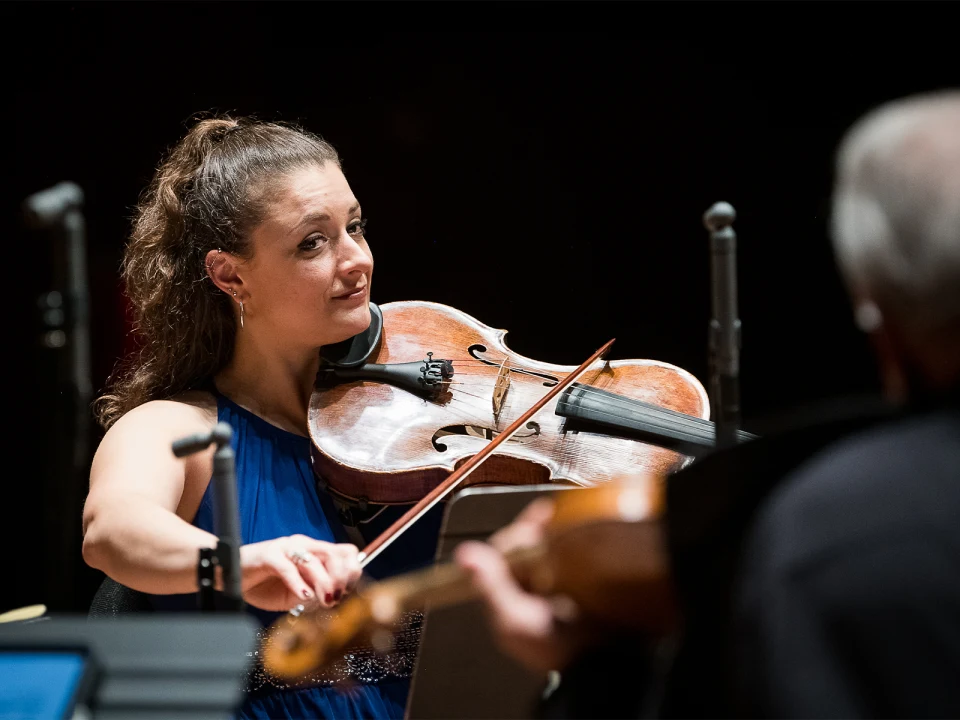 A woman in a blue dress plays the violin, looking at a man playing another string instrument. They are on stage, surrounded by microphones.