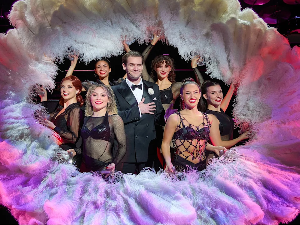 A man in a tuxedo stands surrounded by six women in black and purple dance costumes, framed by large white feather fans on a stage.