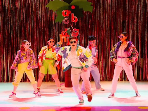 Five people in colorful clothes and sunglasses perform on stage in front of a shiny curtain and a set shaped like a tree with letters hanging from it.