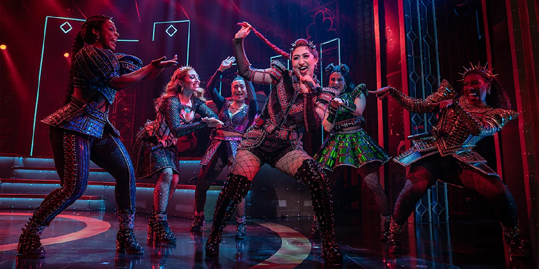 Six women in colorful, elaborate costumes perform energetically on stage under dramatic lighting, with one singer at the center holding a microphone.