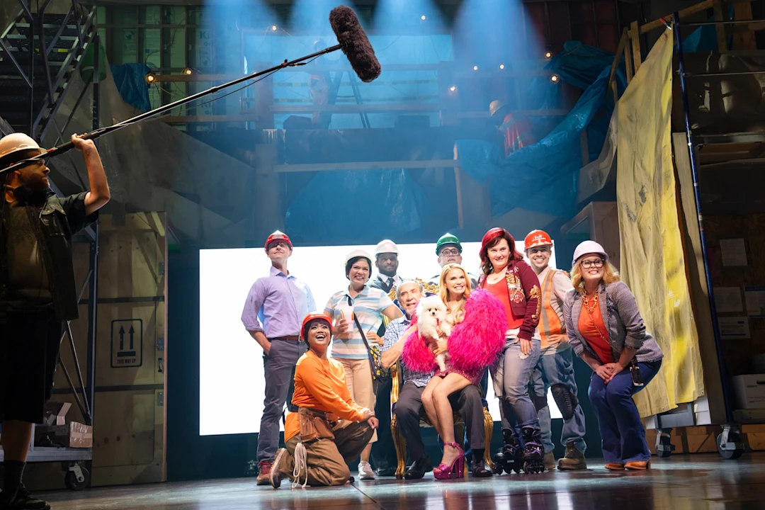 A group of people in colorful costumes and hard hats pose on a stage set, with a boom microphone visible above them.