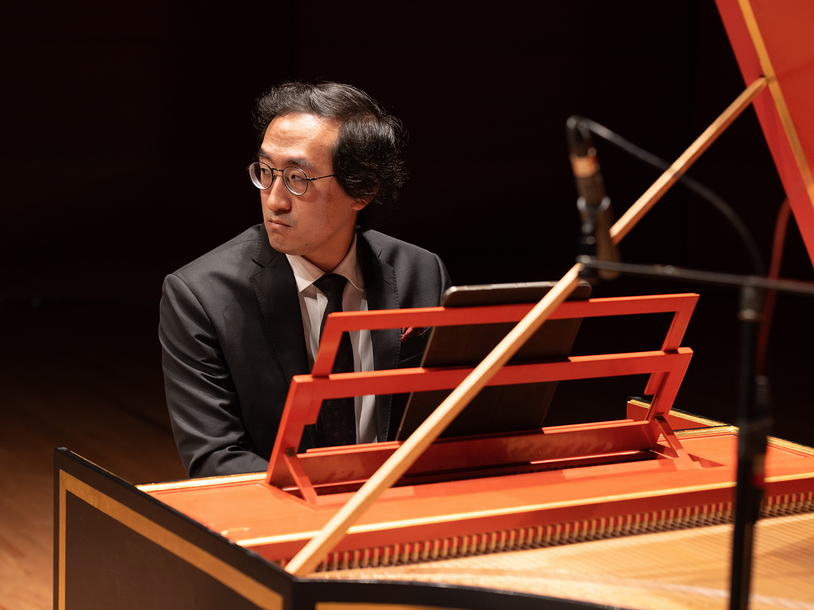 A man in a suit and glasses sits at a harpsichord on stage, looking to the side. A microphone is positioned nearby.