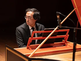 A man in a suit and glasses sits at a harpsichord on stage, looking to the side. A microphone is positioned nearby.