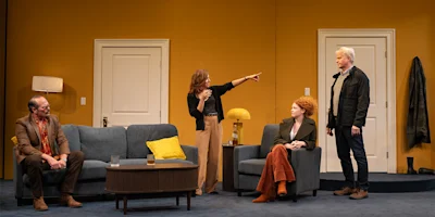 Four adults are in a modern living room; one woman stands and points while the others, two seated and one standing, listen. The setting features yellow walls and contemporary furniture.