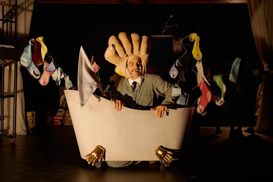 A person in a suit and large hand-shaped hat sits in a bathtub on wheels, decorated with colorful socks and gold hand-shaped feet, on a dimly lit stage.