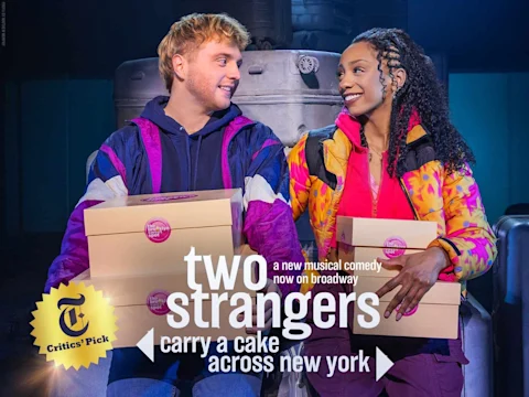 Two people sit side by side, each holding stacked bakery boxes, looking at each other and smiling. The background is dimly lit with industrial containers.