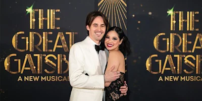 Two people dressed formally pose and smile together in front of a backdrop that reads "THE GREAT GATSBY: A NEW MUSICAL" with gold and black art deco design elements.