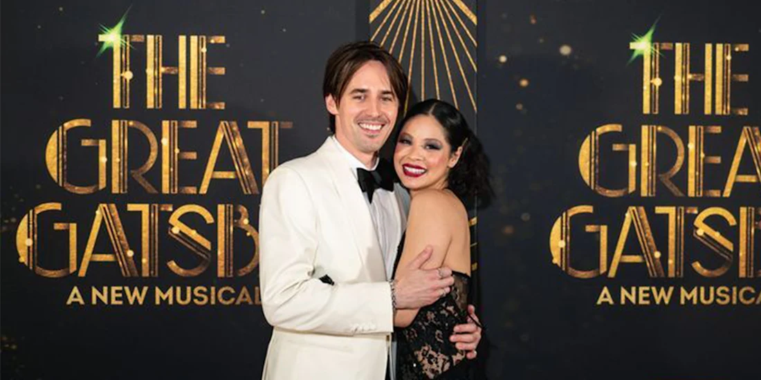 Two people dressed formally pose and smile together in front of a backdrop that reads "THE GREAT GATSBY: A NEW MUSICAL" with gold and black art deco design elements.