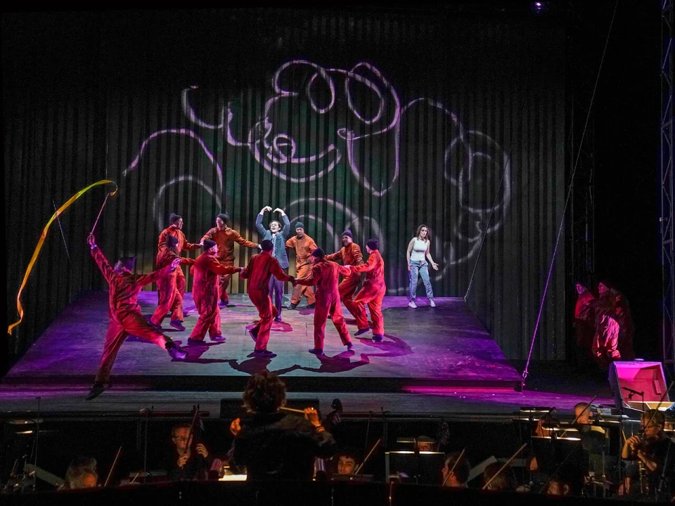 A group of performers in red costumes dance on stage with light trails above, while musicians play in the orchestra pit below. A person in casual wear stands in the background.