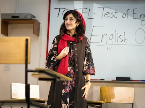 A woman stands in a classroom beside empty chairs, in front of a whiteboard with "TOEFL - Test of English" written on it.