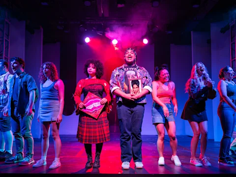 A group of actors perform on stage under colorful lights. The center actors hold a record and a framed photo. The cast stands in a line, singing or speaking with expressive faces.