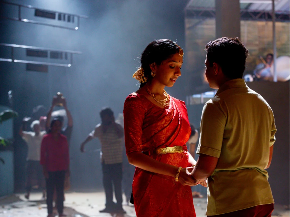 Two people are standing closely, holding hands, and looking at each other. The woman is dressed in a red saree with gold jewelry, while the man is in a green shirt. Several people are in the background.