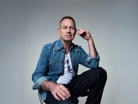 Jason Donovan in a denim jacket and white shirt sits on a chair against a plain, light grey background, resting his hand on his face and looking at the camera.