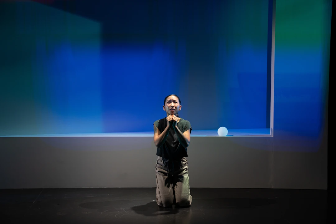A person kneels on a dark stage with hands clasped, looking upward, against a blue-lit abstract background.