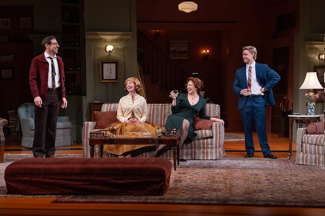 Four adults in vintage-style clothing are gathered in a warmly lit living room set, with two seated on a couch and two standing, engaged in conversation and laughter.