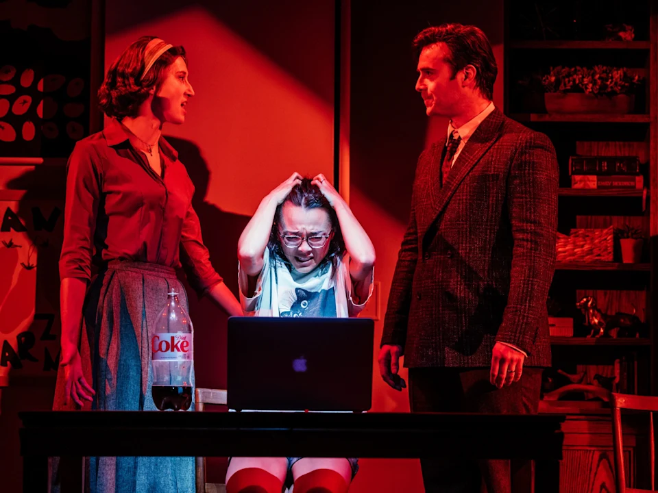 Three people stand around a table with a laptop and a Diet Coke bottle; the person in the middle appears distressed, clutching their head, while the other two look on.
