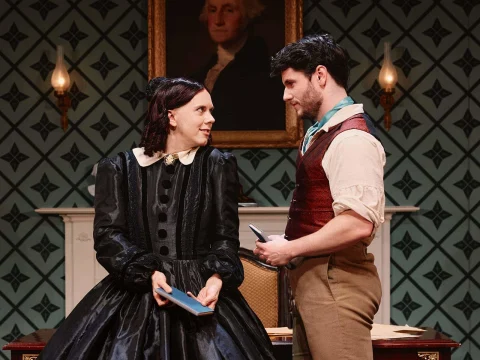 Two people in period costumes, a woman in a black dress and a man in a brown vest, stand indoors holding books, facing each other. A portrait hangs on the patterned wall behind them.