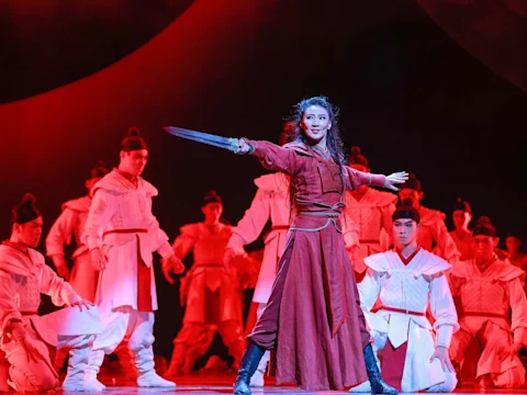 A woman in red wields a sword on stage, surrounded by performers in white costumes, all under dramatic red lighting.