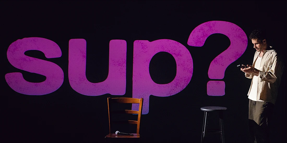 A man looking at his phone on a stage in front of a large, glowing purple “sup?” sign, with a wooden chair and microphone in the foreground.