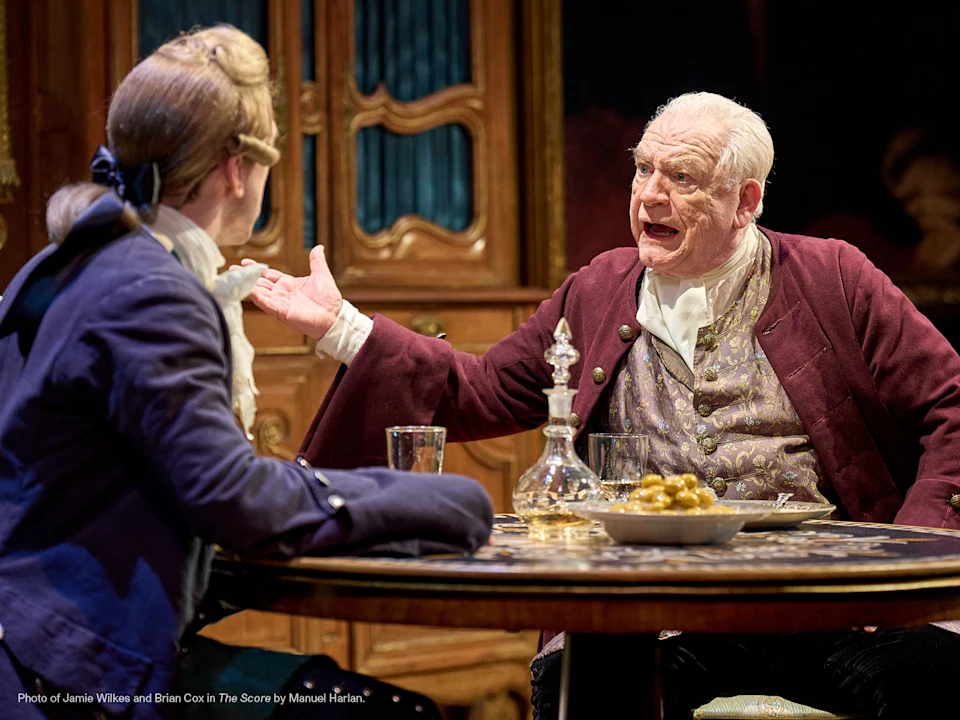 Two men in 18th-century costumes sit at a table with a decanter and glasses; one gestures animatedly while speaking to the other.