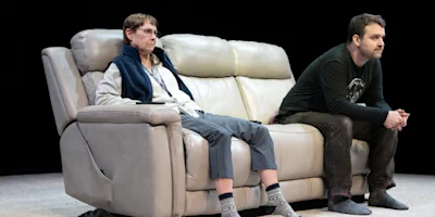 Two people sit on a beige recliner sofa, both facing forward with serious expressions, in a dimly lit setting.