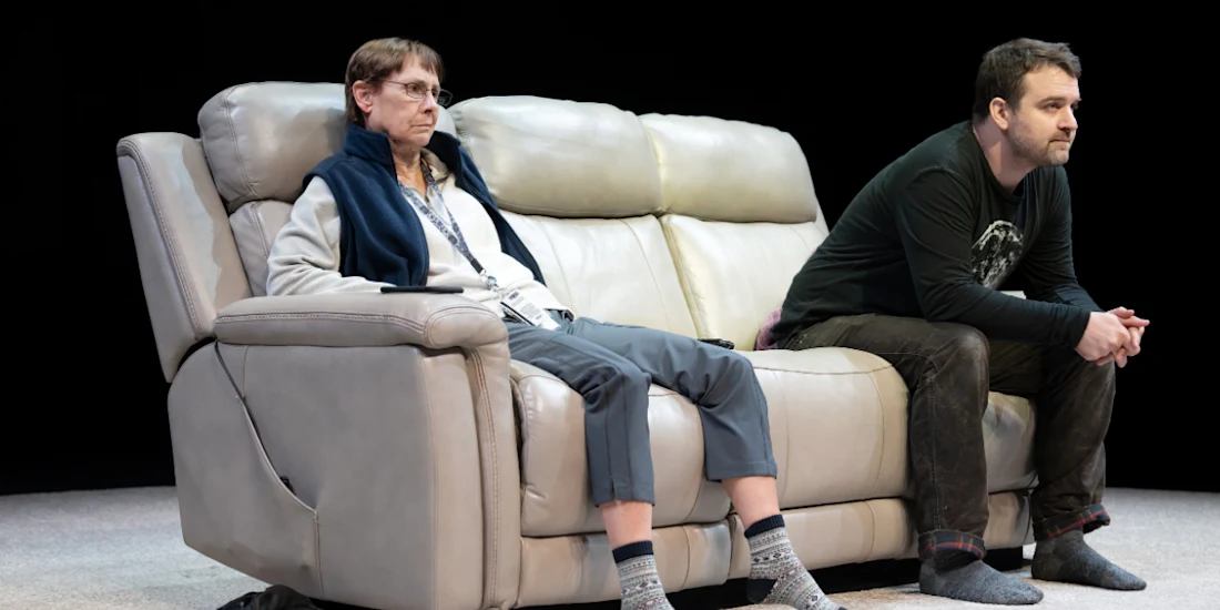 Two people sit on a beige recliner sofa, both facing forward with serious expressions, in a dimly lit setting.