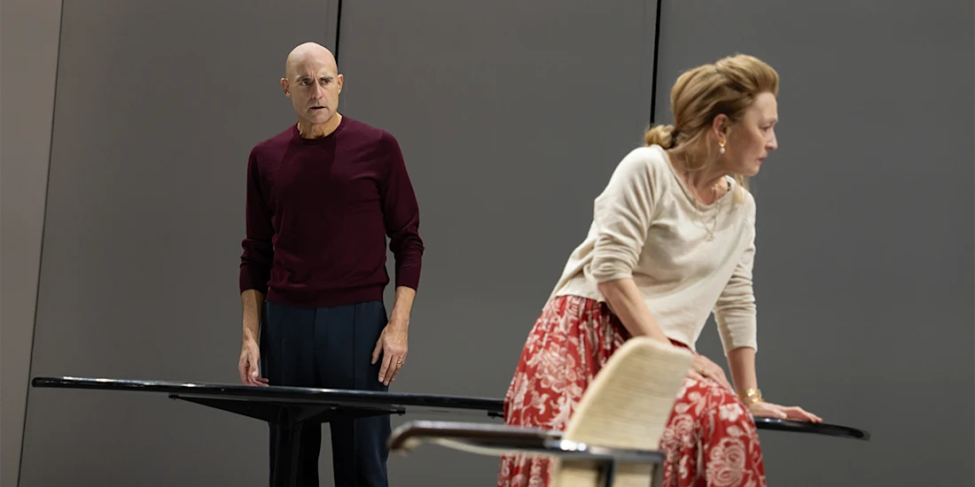 A man stands by a table looking at a woman who sits on the edge of a chair, turned slightly away from him, against a plain background.