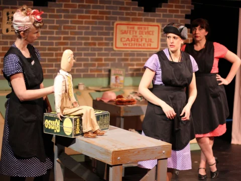 Three women in costumes and aprons are on stage. One woman is holding a puppet, and there is a sign in the background that reads "The best safety device is a careful worker".