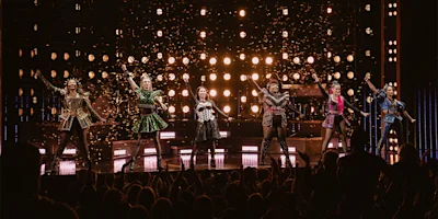 Six performers in colorful, historical costumes stand on stage under bright lights, confetti falling, as an audience applauds in the foreground.