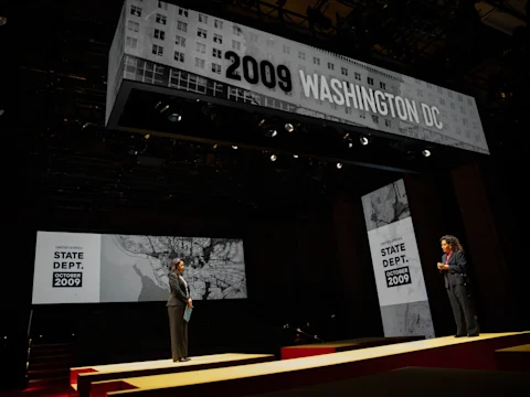 Two people stand on a stage with large digital screens displaying "2009 Washington DC" and "U.S. State Dept. October 2009" with black-and-white city and map visuals.